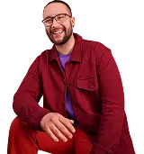 Ein junger Mann mit Brille und Bart sitzt entspannt und lacht in die Kamera. Er trägt eine weinrote Jacke, eine rote Hose und ein lilafarbenes Shirt.