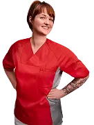 Eine junge Frau in rot-grauer Berufskleidung steht lächelnd mit den Händen in der Hüfte und blickt zur Seite. Sie hat kurze braune Haare und ein Tattoo am rechten Unterarm.